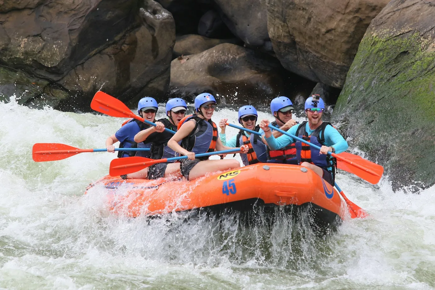 Turuncu bir rafting botunda kask ve can yeleği giymiş bir grup, kayalar arasındaki köpüklü suda kürek çekiyor