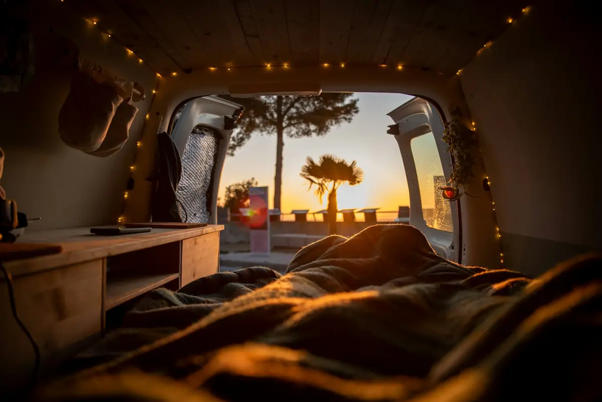Innenraum eines Campers bei Sonnenuntergang mit offenen Hecktüren; hinter dem Bett mit Decke eine Palme und das Meer
