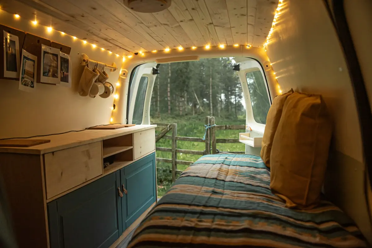 Innenraum eines Campers mit Holzdecke: rechts ein Bett mit gestreifter Decke, links ein Küchenblock mit petrolfarbenen Türen, durch die offene Hecktür ist Wald zu sehen
