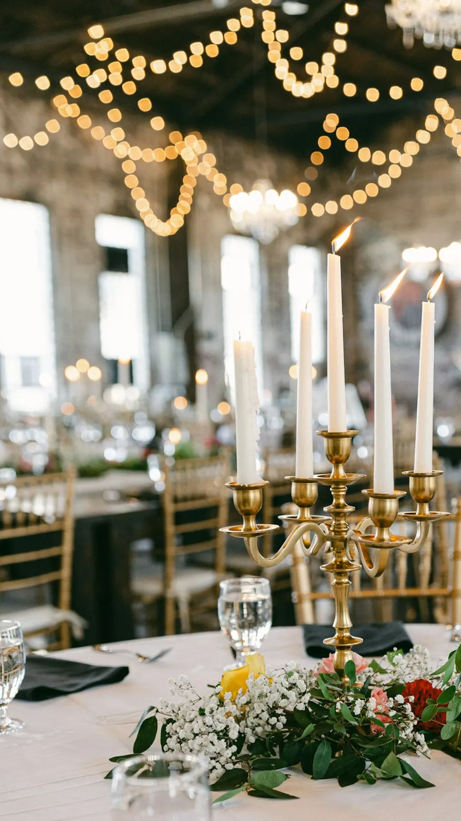 Tall candles burning in a brass candelabra on a round table with greenery and white flowers, string lights in the background