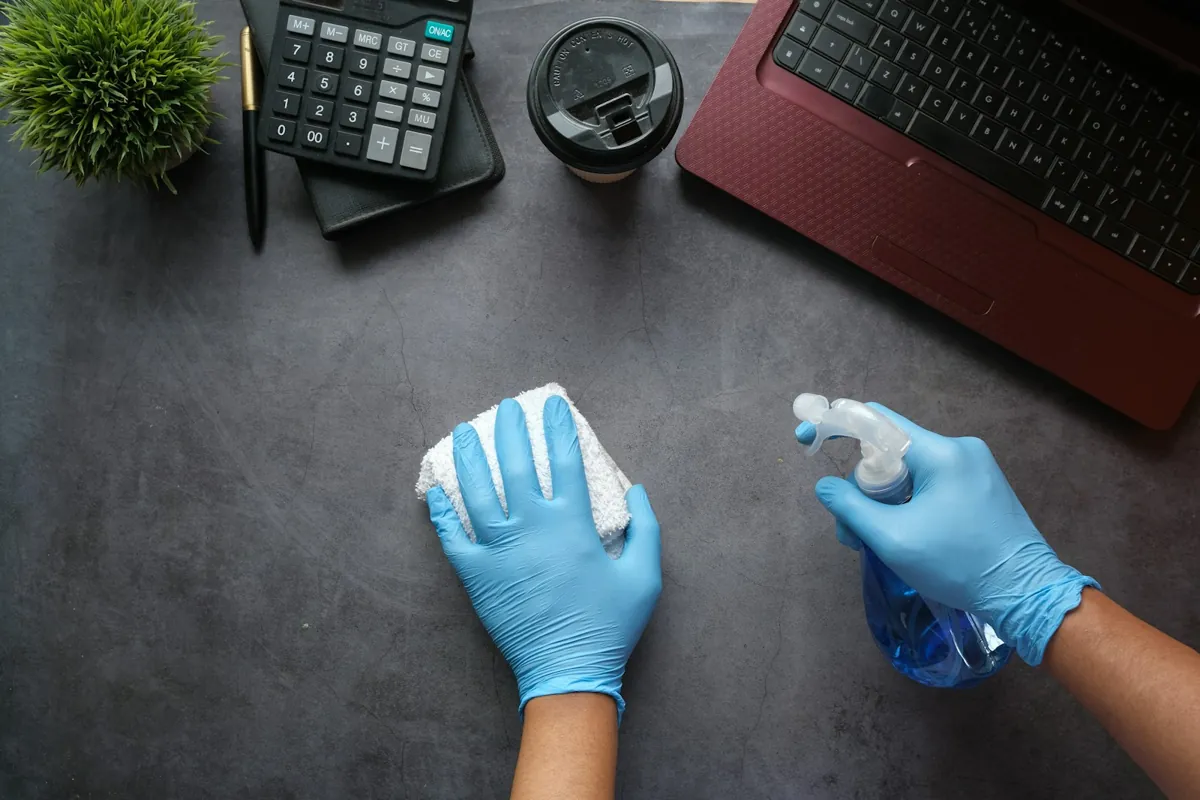 Hände in Handschuhen wischen einen Bürotisch mit Spray und Tuch